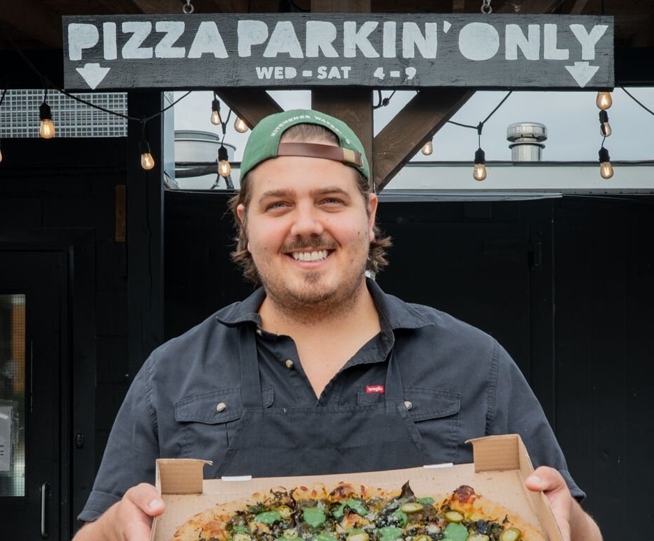 Chef Tim Borys standing in front of Back of House Pizza’s pickup door beneath the “Pizza Parkin’ Only” sign, holding an open box with a fresh feature pizza partly visible.